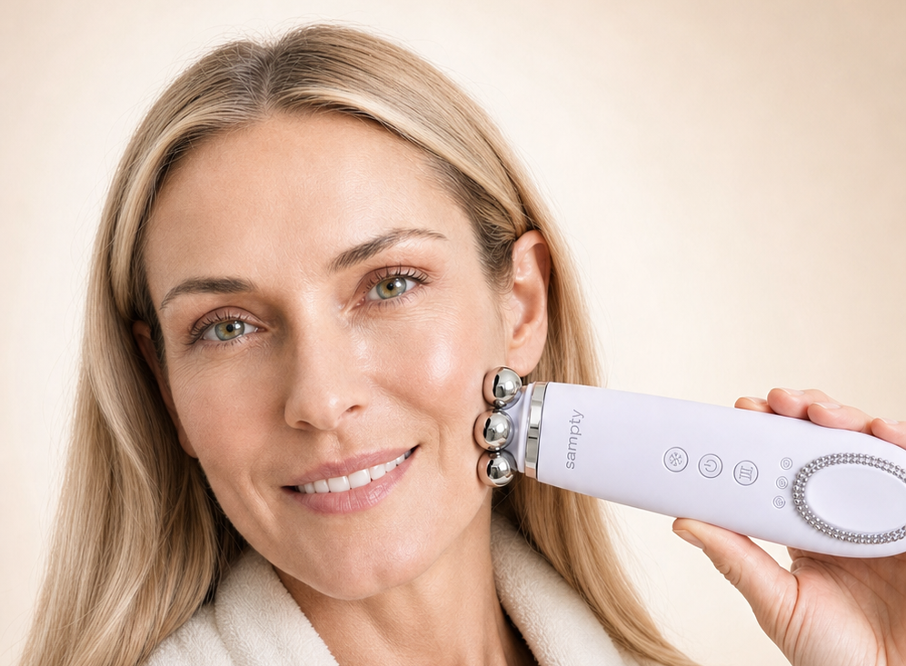 A smiling blonde woman in a cream bathrobe holds a white and silver facial skincare device against her cheek, set against a soft beige background.