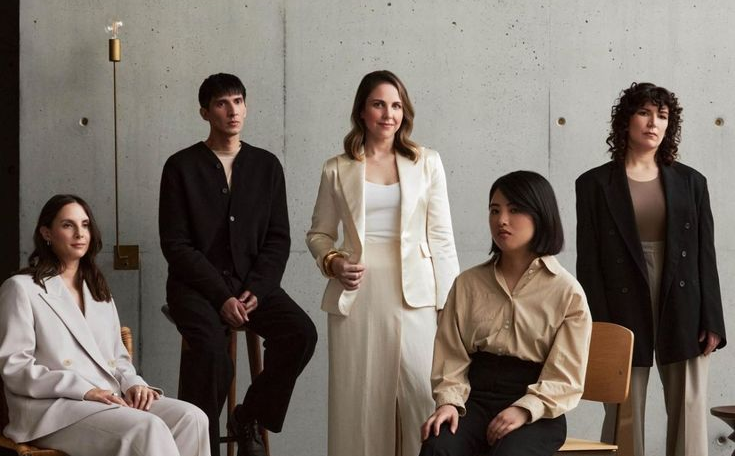 Five fashion models in stylish outfits pose together in a minimalist studio setting with a concrete wall background.